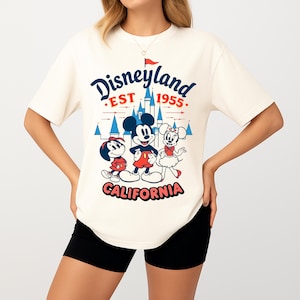 Puede incluir: Camiseta color crema con el logo de Disneyland y Mickey, Minnie y Donald. El gr&aacute;fico incluye el texto "Disneyland EST 1955 CALIFORNIA" en azul y rojo.
