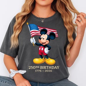 Puede incluir: Camiseta gris oscuro con Mickey Mouse sosteniendo una bandera estadounidense. Mickey lleva pantalones cortos rojos, zapatos amarillos y guantes blancos. El texto "250th BIRTHDAY 1776-2026" est&aacute; impreso debajo. La camiseta es de corte holgado.