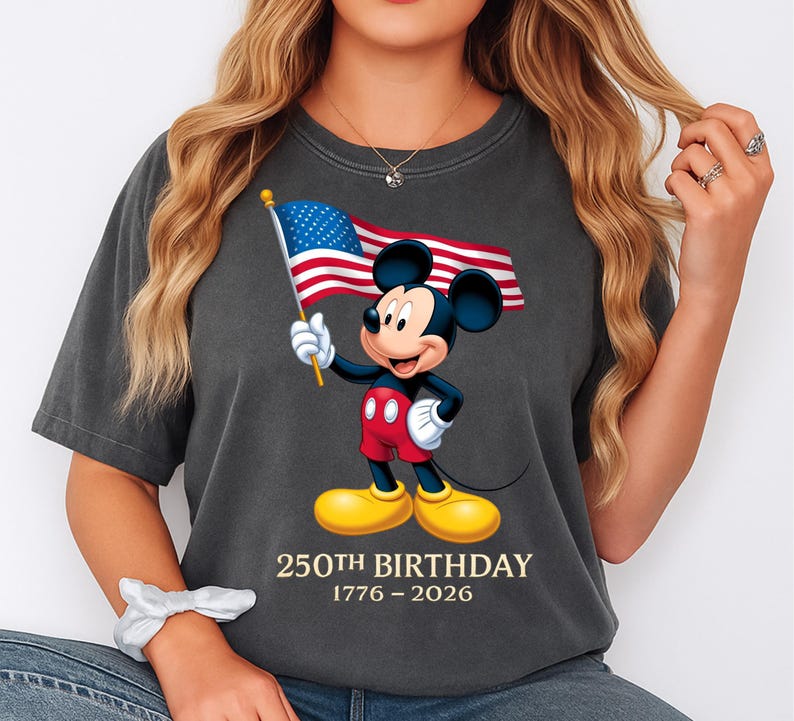 Puede incluir: Camiseta gris oscuro con Mickey Mouse sosteniendo una bandera estadounidense. Mickey lleva pantalones cortos rojos, zapatos amarillos y guantes blancos. El texto "250th BIRTHDAY 1776-2026" est&aacute; impreso debajo. La camiseta es de corte holgado.