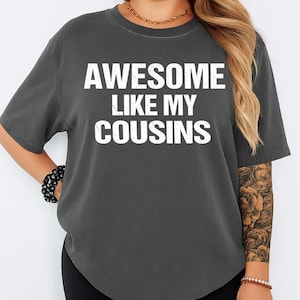 K&ouml;nnte beinhalten: Dunkelgraues T-Shirt mit dem wei&szlig;en Text "AWESOME LIKE MY COUSINS". Das Shirt hat kurze &Auml;rmel und einen Rundhalsausschnitt. Das Material wirkt weich.