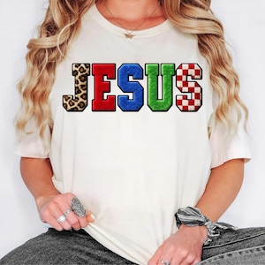 May include: Cream-colored t-shirt with the word "JESUS" in bold, colorful letters. The "J" has a leopard print, the "E" is red, the "S" is blue, the second "S" is green, and the "U" has a red and white checkered pattern.