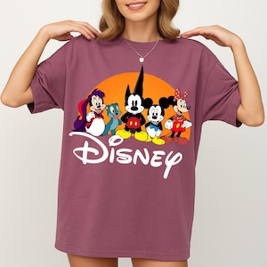 Pu&ograve; includere: T-shirt color malva con grafica Disney. La grafica include Topolino, Minnie e altri personaggi Disney davanti a un sole arancione. La parola "Disney" &egrave; scritta in bianco.