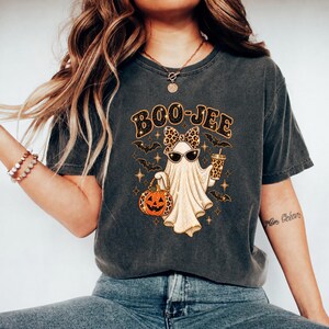 May include: A charcoal gray short-sleeve t-shirt featuring a graphic of a ghost wearing a leopard print bow and sunglasses. The ghost holds a pumpkin and a leopard print cup. The text BOO-JEE is printed above the ghost with small bats and stars.