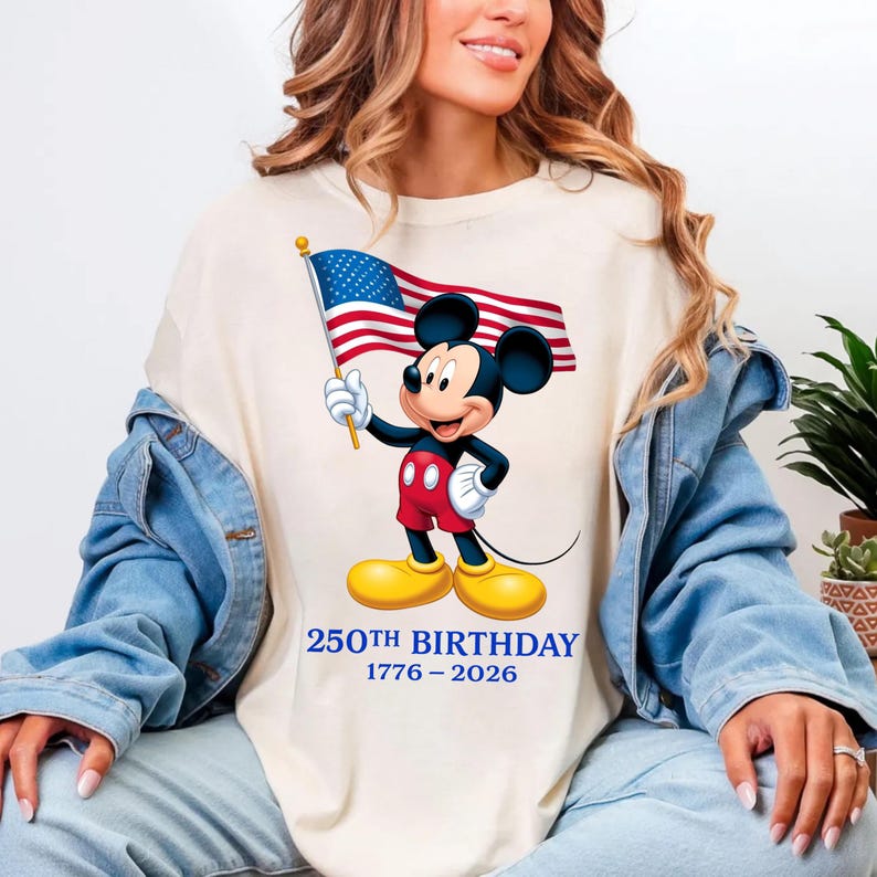 Puede incluir: Camiseta color crema con Mickey Mouse sosteniendo una bandera estadounidense. Mickey lleva pantalones cortos rojos, una chaqueta negra, zapatos amarillos y guantes blancos. El texto "250TH BIRTHAY 1776-2026" est&aacute; impreso en azul debajo.