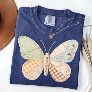 May include: A navy blue Comfort Colors t-shirt featuring a patchwork butterfly design. The butterfly has wings with various patterns, including polka dots, gingham, and floral prints. A gold necklace with a pendant rests on the shirt.