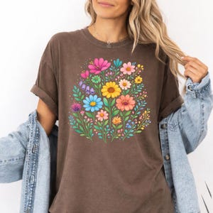 May include: A brown t-shirt featuring a vibrant floral design. The graphic showcases a bouquet of colorful flowers, including pink, yellow, blue, and orange blooms, surrounded by green leaves. The shirt has rolled-up sleeves and is paired with a denim jacket.
