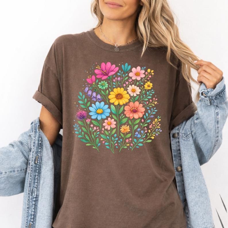 May include: A brown t-shirt featuring a vibrant floral design. The graphic showcases a bouquet of colorful flowers, including pink, yellow, blue, and orange blooms, surrounded by green leaves. The shirt has rolled-up sleeves and is paired with a denim jacket.