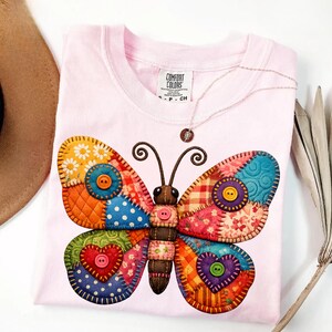 May include: A light pink t-shirt featuring a colorful butterfly design. The butterfly is made of patchwork fabric with various patterns and colors, including orange, blue, and red. The shirt has a crew neck and short sleeves.