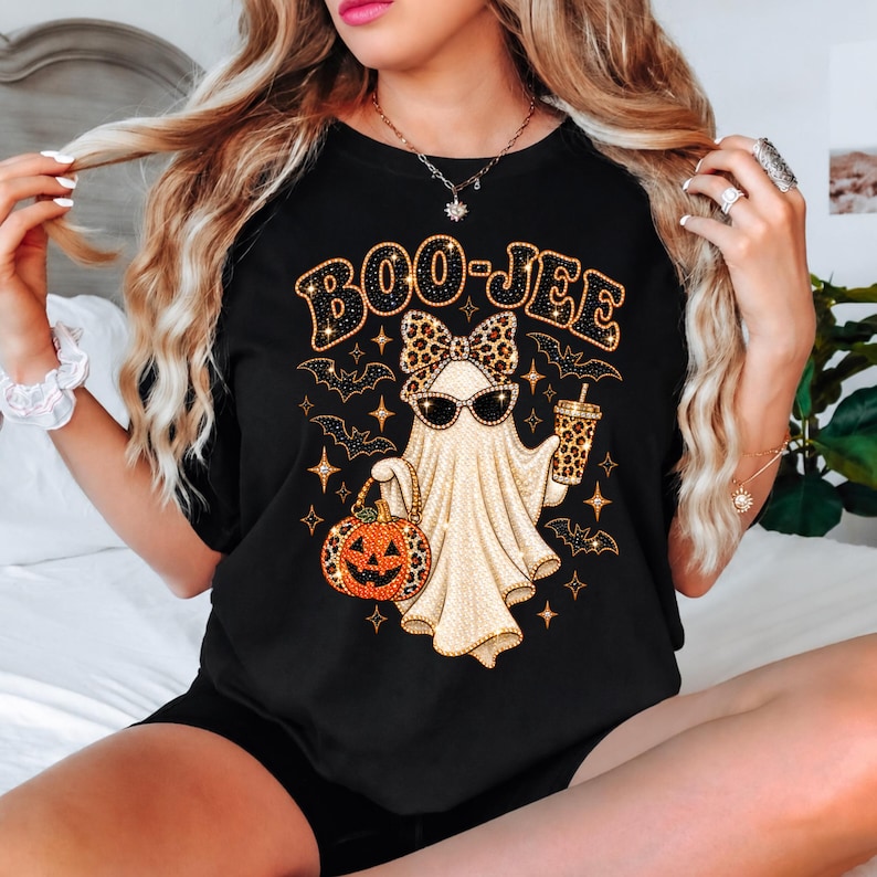 May include: A black t-shirt featuring a graphic of a ghost wearing sunglasses and a leopard print bow. The ghost holds a leopard print cup and a pumpkin with a leopard print handle. The text BOO-JEE is printed above in a glittery gold font.