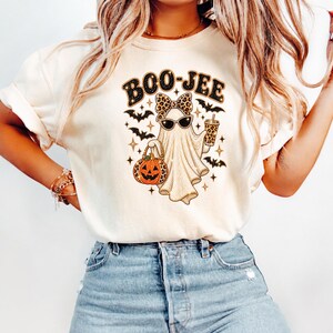 May include: A cream-colored t-shirt featuring a graphic of a ghost wearing sunglasses and a leopard print bow. The ghost holds a jack-o'-lantern and a drink cup, surrounded by bats and stars. The text BOO-JEE is printed above the ghost.