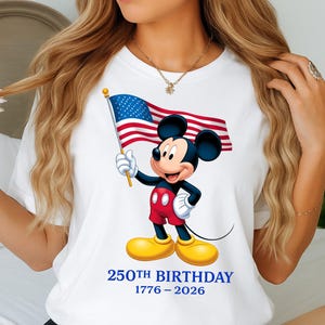 Puede incluir: Camiseta blanca con Mickey Mouse sosteniendo una bandera estadounidense. Mickey lleva pantalones cortos rojos, zapatos amarillos y guantes blancos. El texto "250TH BIRTHAY 1776-2026" est&aacute; impreso en azul debajo.
