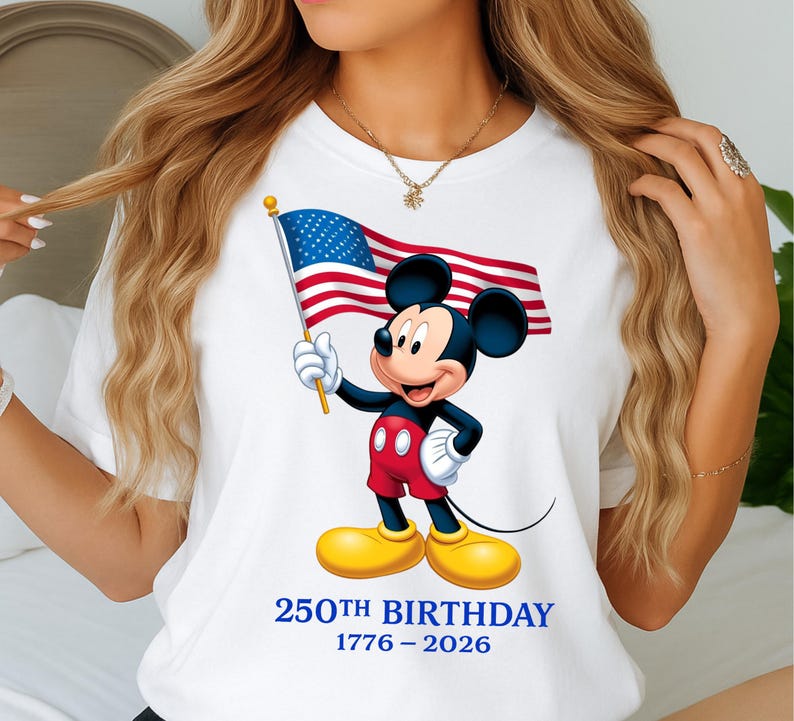 Puede incluir: Camiseta blanca con Mickey Mouse sosteniendo una bandera estadounidense. Mickey lleva pantalones cortos rojos, zapatos amarillos y guantes blancos. El texto "250TH BIRTHAY 1776-2026" est&aacute; impreso en azul debajo.