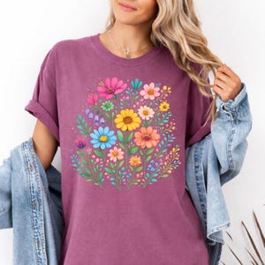 May include: A short-sleeved, mauve t-shirt featuring a vibrant floral design. The graphic showcases a bouquet of colorful flowers, including pink, yellow, blue, and orange blooms, surrounded by green leaves. The shirt has a relaxed fit.