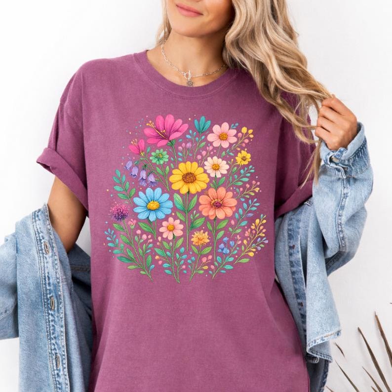 May include: A short-sleeved, mauve t-shirt featuring a vibrant floral design. The graphic showcases a bouquet of colorful flowers, including pink, yellow, blue, and orange blooms, surrounded by green leaves. The shirt has a relaxed fit.