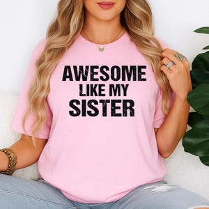 K&ouml;nnte beinhalten: Hellrosa T-Shirt mit dem Text "AWESOME LIKE MY SISTER" in fetten schwarzen Buchstaben. Das Shirt hat einen Rundhalsausschnitt und scheint aus einem weichen Material zu sein. Das Thema ist Freizeitkleidung.
