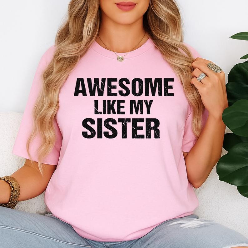 K&ouml;nnte beinhalten: Hellrosa T-Shirt mit dem Text "AWESOME LIKE MY SISTER" in fetten schwarzen Buchstaben. Das Shirt hat einen Rundhalsausschnitt und scheint aus einem weichen Material zu sein. Das Thema ist Freizeitkleidung.