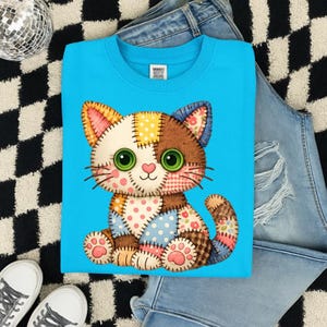 May include: A light blue sweatshirt featuring a patchwork cat design. The cat has green eyes, a pink nose, and is made of various patterned fabrics. The sweatshirt is displayed with a pair of jeans and sneakers on a black and white checkered surface.