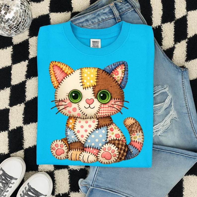 May include: A light blue sweatshirt featuring a patchwork cat design. The cat has green eyes, a pink nose, and is made of various patterned fabrics. The sweatshirt is displayed with a pair of jeans and sneakers on a black and white checkered surface.