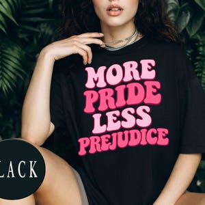 May include: A black short-sleeved t-shirt featuring the text "MORE PRIDE LESS PREJUDICE" printed in a bold, wavy, pink font across the chest. The garment has a crew neckline and a relaxed fit, with the word "BLACK" visible in a circular badge.