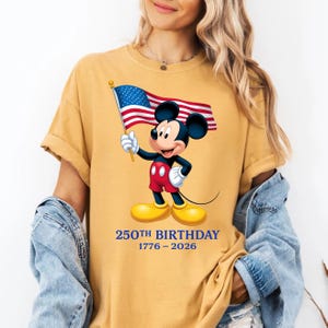 Puede incluir: Camiseta amarilla mostaza con un dibujo de Mickey Mouse sosteniendo una bandera estadounidense. Mickey lleva pantalones cortos rojos, guantes blancos y zapatos amarillos. El texto "250TH BIRTHDAY 1776-2026" est&aacute; impreso debajo.