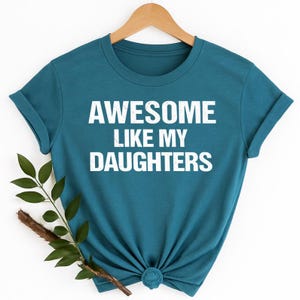 K&ouml;nnte beinhalten: T&uuml;rkisfarbenes T-Shirt mit dem wei&szlig;en Text "AWESOME LIKE MY DAUGHTERS". Das T-Shirt h&auml;ngt an einem Holzb&uuml;gel. Ein Zweig mit gr&uuml;nen Bl&auml;ttern und einem braunen Stiel befindet sich in der linken unteren Ecke.