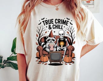 Könnte beinhalten: Ein cremefarbenes T-Shirt mit einer Grafik eines Waschbären mit Hexenhut und eines Skeletts, die auf einem braunen Sofa sitzen. Der Text TRUE CRIME & CHILL steht darüber. Die Szene zeigt einen Fernseher, Popcorn, Kürbisse und Bäume.