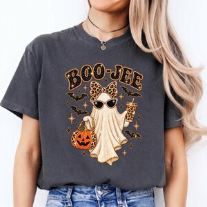 May include: A charcoal gray t-shirt featuring a graphic of a ghost wearing a leopard print bow and sunglasses, holding a pumpkin bucket and a drink. The text BOO-JEE is printed above the ghost, surrounded by small bats and sparkling star accents.