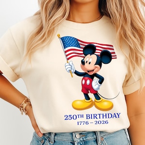 Puede incluir: Camiseta color crema con un gr&aacute;fico de Mickey Mouse sosteniendo una bandera estadounidense. Mickey lleva pantalones cortos rojos, guantes negros y zapatos amarillos. El texto "250th BIRTHDAY 1776-2026" est&aacute; impreso en azul debajo.