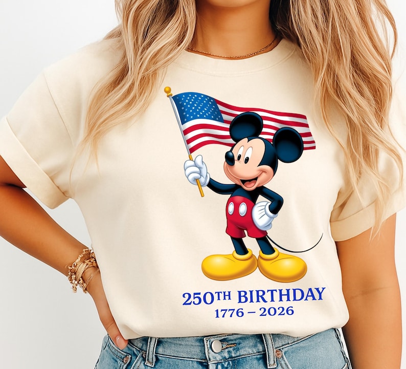 Puede incluir: Camiseta color crema con un gr&aacute;fico de Mickey Mouse sosteniendo una bandera estadounidense. Mickey lleva pantalones cortos rojos, guantes negros y zapatos amarillos. El texto "250th BIRTHDAY 1776-2026" est&aacute; impreso en azul debajo.