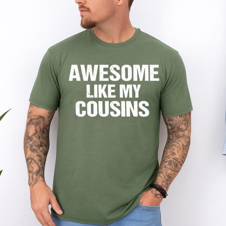 K&ouml;nnte beinhalten: Olivgr&uuml;nes T-Shirt mit dem wei&szlig;en Text "AWESOME LIKE MY COUSINS". Das T-Shirt hat kurze &Auml;rmel und einen Rundhalsausschnitt. Die Person, die das T-Shirt tr&auml;gt, hat T&auml;towierungen auf den Armen.