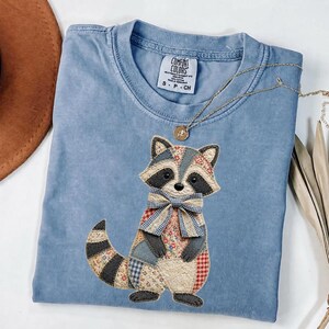 May include: A light blue Comfort Colors t-shirt featuring a patchwork raccoon design with a bow tie. The raccoon has black and gray accents. A gold necklace with a pendant is draped over the shirt. The shirt is size small.