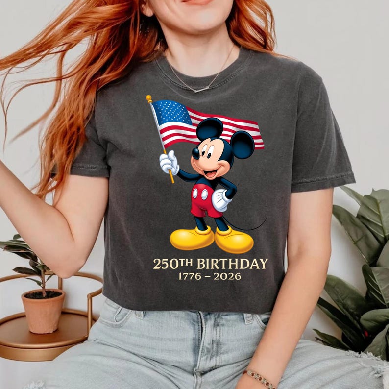 Puede incluir: Camiseta gris oscuro con Mickey Mouse sosteniendo una bandera estadounidense. Mickey lleva pantalones cortos rojos, guantes blancos y zapatos amarillos. El texto "250TH BIRTHDAY 1776-2026" est&aacute; impreso debajo.