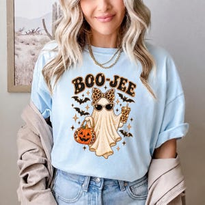 May include: A light blue t-shirt featuring a graphic of a ghost wearing leopard print sunglasses and a matching bow. The ghost holds a leopard print pumpkin bucket and a drink cup, surrounded by black bats and stars with the text BOO-JEE.