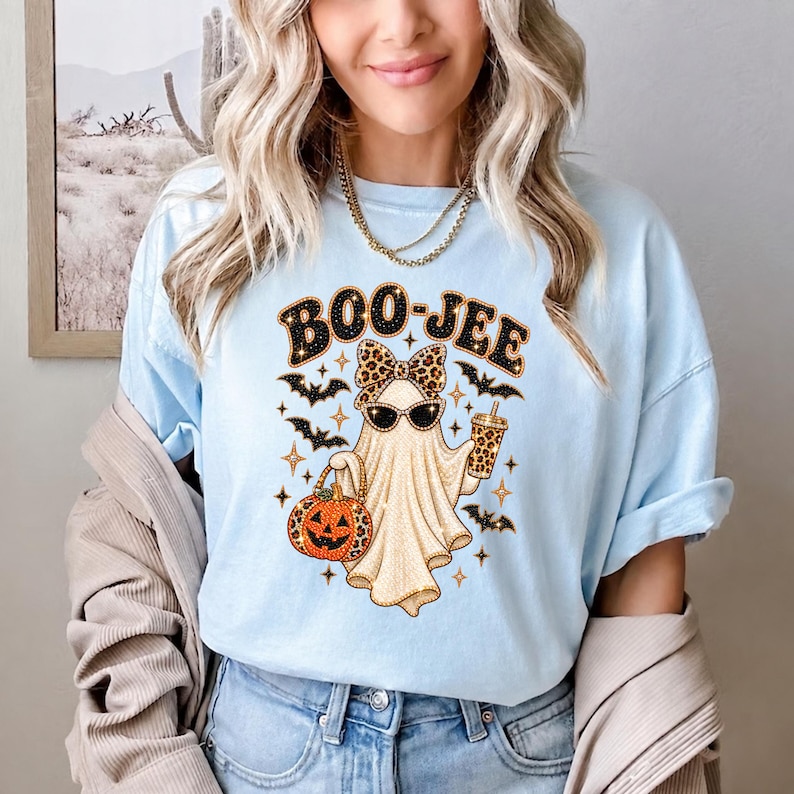 May include: A light blue t-shirt featuring a graphic of a ghost wearing leopard print sunglasses and a matching bow. The ghost holds a leopard print pumpkin bucket and a drink cup, surrounded by black bats and stars with the text BOO-JEE.