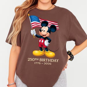 Puede incluir: Camiseta marr&oacute;n con Mickey Mouse sosteniendo una bandera estadounidense. Mickey lleva pantalones cortos rojos, guantes blancos y zapatos amarillos. El texto "250th BIRTHDAY 1776-2026" est&aacute; impreso debajo.