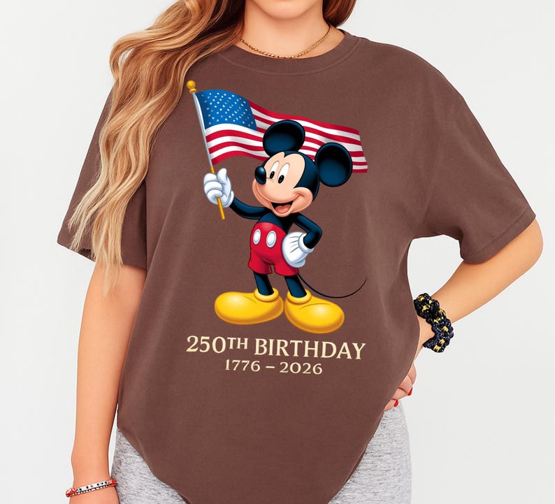 Puede incluir: Camiseta marr&oacute;n con Mickey Mouse sosteniendo una bandera estadounidense. Mickey lleva pantalones cortos rojos, guantes blancos y zapatos amarillos. El texto "250th BIRTHDAY 1776-2026" est&aacute; impreso debajo.