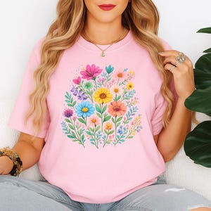 May include: A light pink t-shirt featuring a vibrant floral design. The design showcases a bouquet of colorful flowers, including pink, yellow, blue, and orange blooms, surrounded by green leaves. The shirt is made of soft material.