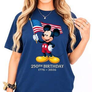 Puede incluir: Camiseta azul marino con Mickey Mouse sosteniendo una peque&ntilde;a bandera estadounidense. Mickey lleva pantalones cortos rojos, guantes blancos y zapatos amarillos. El texto "250th Birthday 1776-2026" est&aacute; impreso debajo.