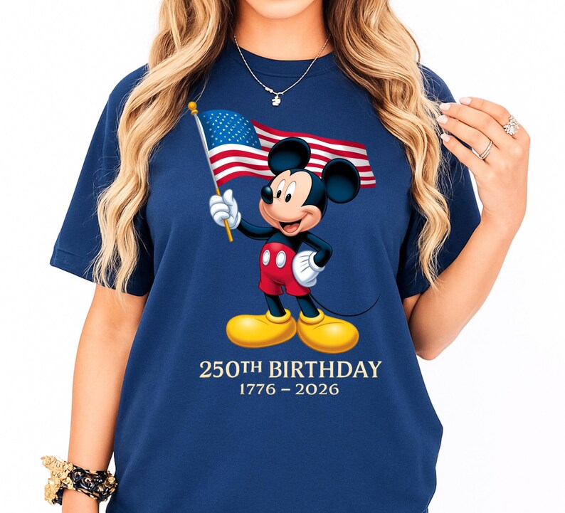 Puede incluir: Camiseta azul marino con Mickey Mouse sosteniendo una peque&ntilde;a bandera estadounidense. Mickey lleva pantalones cortos rojos, guantes blancos y zapatos amarillos. El texto "250th Birthday 1776-2026" est&aacute; impreso debajo.