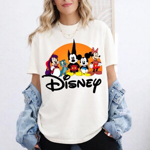 Pu&ograve; includere: T-shirt color crema con grafica Disney. La grafica include Topolino, Minnie e altri personaggi Disney davanti a un sole arancione. La parola "Disney" &egrave; scritta in nero sotto i personaggi.