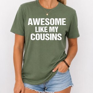 K&ouml;nnte beinhalten: Ein olivgr&uuml;nes T-Shirt mit dem Text "AWESOME LIKE MY COUSINS" in wei&szlig;en Blockbuchstaben. Das kurz&auml;rmelige T-Shirt scheint aus einem weichen Material zu sein. Das T-Shirt wird von einer Person getragen.