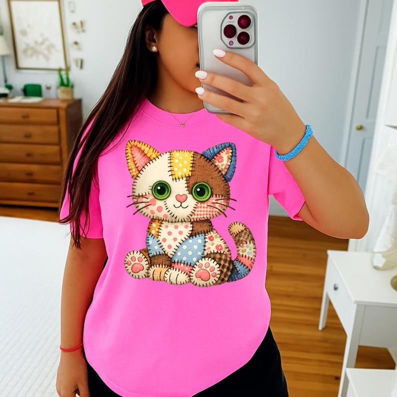 May include: A bright pink t-shirt featuring a colorful patchwork cat design. The cat has large green eyes, a pink nose, and a variety of patterned patches in its design. The shirt is a solid pink color.