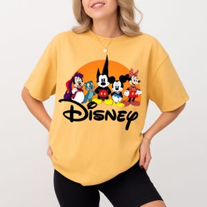 Pu&ograve; includere: T-shirt giallo senape con grafica Disney. La grafica include Topolino, Minnie e altri personaggi Disney in un design colorato. La parola "Disney" &egrave; scritta in nero sotto i personaggi.