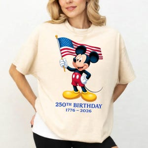 Puede incluir: Camiseta color crema con Mickey Mouse sosteniendo una bandera estadounidense. Mickey lleva pantalones cortos rojos, zapatos amarillos y guantes blancos. El texto "250TH BIRTHAY 1776-2026" est&aacute; impreso debajo en azul.