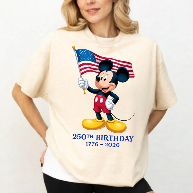 Puede incluir: Camiseta color crema con Mickey Mouse sosteniendo una bandera estadounidense. Mickey lleva pantalones cortos rojos, zapatos amarillos y guantes blancos. El texto "250TH BIRTHAY 1776-2026" est&aacute; impreso debajo en azul.