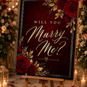 Könnte beinhalten: Burgunderfarbenes Schild auf einer Holzstaffelei mit goldener Aufschrift "Will You Marry Me?". Das Schild ist mit roten Rosen und goldenen Blättern verziert. Unter den Worten ist ein Herz gezeichnet. Der Hintergrund ist verschwommen mit Blumen und Kerzen.
