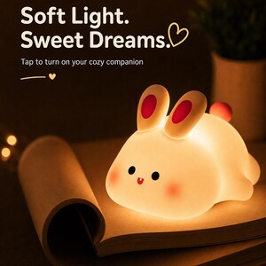 May include: A soft, glowing bunny-shaped night light with red-tipped ears and rosy cheeks. The text "Soft Light. Sweet Dreams." and "Tap to turn on your cozy companion" are displayed. The light rests on an open book.