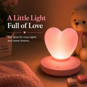 Puede incluir: Una lámpara de noche rosa en forma de corazón con el texto "A Little Light Full of Love". La lámpara se encuentra sobre una base redonda con un botón de encendido. También se ven un osito de peluche y decoraciones en forma de corazón.