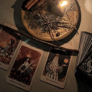 Puede incluir: Una escena tenue con cartas del tarot, una vela encendida y un cuenco de lat&oacute;n con hierbas y palitos de canela. Las cartas muestran ilustraciones y texto, incluyendo "QUEEN OF SWORDS" y "THE MAGICIAN". Una varita de madera descansa sobre las cartas.