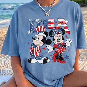 May include: Blue jean t-shirt featuring Mickey and Minnie Mouse in patriotic attire with the text "USA". Mickey wears a hat and suit with red, white, and blue stripes and stars. Minnie wears a red and white polka dot skirt and bow.